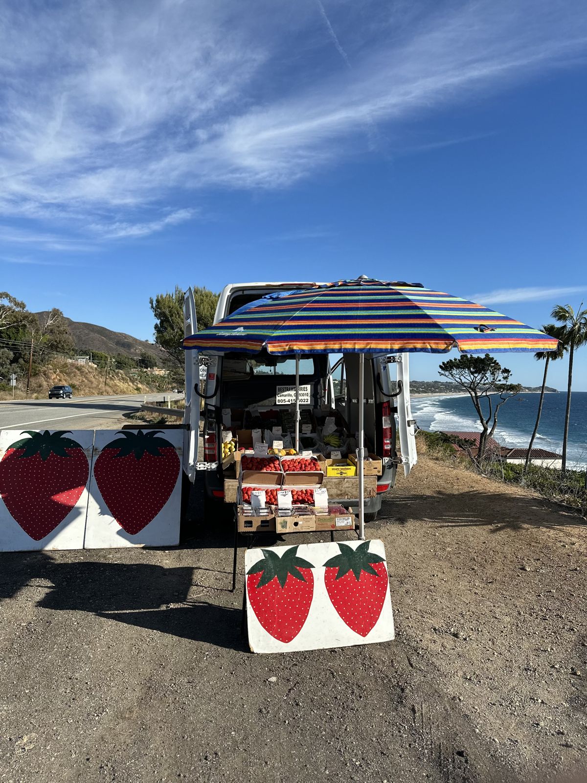 The Bestrawberries van and fruit stand under a striped umbrella, with painted strawberry signs and the ocean behind